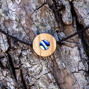 Peut inclure: Un collier pendentif en bois avec un pendentif rond en bois de couleur claire. Le pendentif présente un motif marbré bleu et blanc au centre. Le cordon du collier est marron foncé et le fond est un écorce d'arbre texturée.