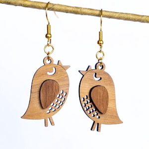 Peut inclure: Une paire de boucles d'oreilles en bois en forme d'oiseau avec des crochets dorés. Les boucles d'oreilles présentent une forme d'oiseau brun clair avec un incrustation brun foncé et de petits détails découpés. Elles sont suspendues à un fil doré sur fond blanc.