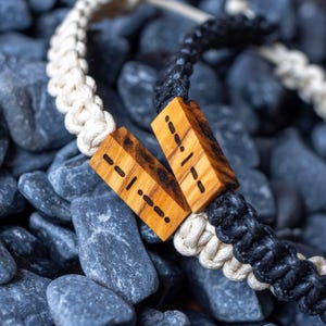 Puede incluir: Dos pulseras de cordón trenzado con cuentas rectangulares de madera grabadas con patrones de código Morse. Una pulsera tiene un cordón de color crema y la otra un cordón negro. Las cuentas de madera muestran una veta natural con marcas marrones.