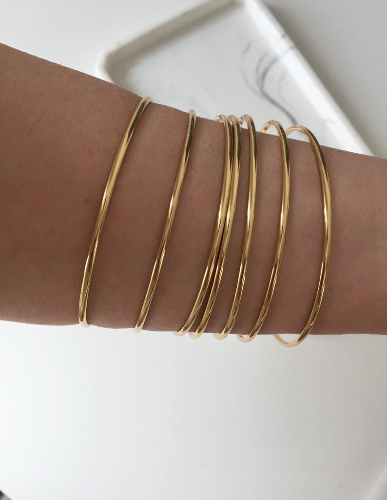 May include: A collection of thin, gold-colored bangles stacked on an arm. The bangles are smooth and shiny, reflecting light. The arm is fair-skinned, and the background is a blurred white surface.