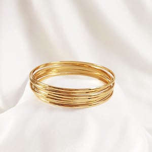 May include: A stack of gold-colored bangles. The bracelets are thin and round, with a smooth, shiny finish. The bangles are resting on a white, silky fabric, creating a simple and elegant display. The jewelry is suitable for everyday wear or special occasions.