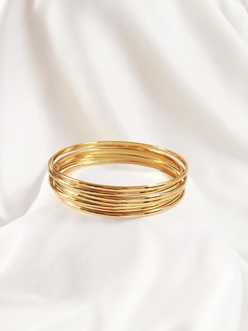 May include: A stack of gold-colored bangles. The bracelets are thin and round, with a smooth, shiny finish. The bangles are resting on a white, silky fabric, creating a simple and elegant display. The jewelry is suitable for everyday wear or special occasions.