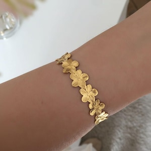 Könnte beinhalten: Ein goldfarbenes Armband mit einer Reihe von blütenförmigen Anhängern. Das Armband besteht aus glänzendem Metall und wird am Handgelenk getragen. Die Blumen sind mit geätzten Linien detailliert, und das Armband scheint ein zartes Schmuckstück zu sein.