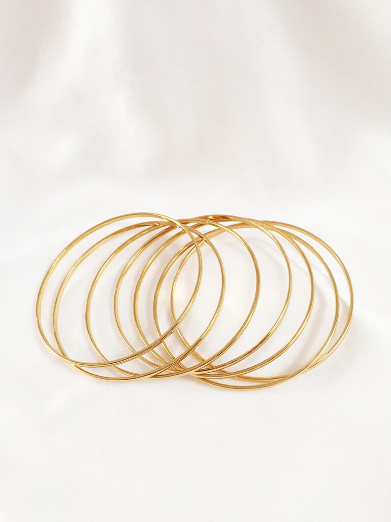 May include: A collection of thin, gold-colored bangles. The bangles are circular and appear to be made of metal. They are arranged in a slightly overlapping pattern against a white background, creating a simple yet elegant aesthetic.