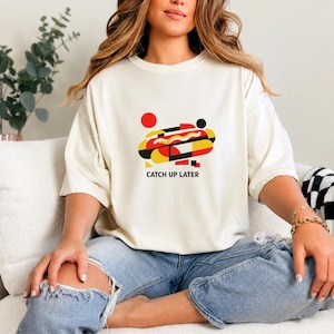 May include: An off-white short-sleeve t-shirt featuring a graphic design of a stylized hot dog in red, yellow, and black geometric shapes. Below the graphic, the text CATCH UP LATER is printed in black capital letters on the front of the shirt.