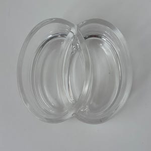 May include: A clear, double-oval shaped glass dish. The dish is divided into two sections, connected by a curved handle. The glass is transparent, allowing light to pass through. The dish is likely used for serving or displaying small items.