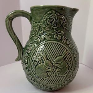 May include: Green ceramic pitcher with a handle and spout. The pitcher features embossed floral and bunny designs. The body of the pitcher has a circular design with two bunnies. The pitcher is a decorative item.