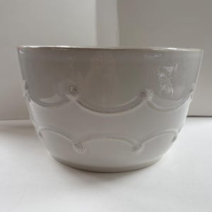 May include: A white ceramic bowl featuring a smooth, glossy finish and a decorative embossed scalloped pattern around the exterior. The design includes small, raised circular details at the points where the scalloped lines meet, creating a textured border.