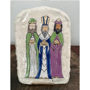 May include: A ceramic plaque featuring three figures, each holding a golden box. The figures are depicted in robes of green, blue, and purple, with beards and crowns. The plaque has a textured, off-white background.