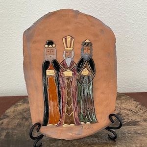 Pode incluir: Uma placa de cor terracota com um relevo dos Três Reis Magos. As figuras são retratadas em vestes coloridas, segurando presentes. A placa é montada em um suporte de metal preto, adequado para exibição.