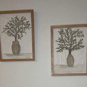 May include: Two framed wall art pieces. Each features a textured, off-white background with a vase holding a tree with green leaves. The frames are made of light-colored wood. The art has a neutral color palette.