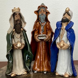 May include: Three ceramic Wise Men figurines. The figures wear robes in green, brown, and blue, with gold accents. Each holds a small, gold-colored vessel. The figures have beards and are wearing crowns or head coverings.