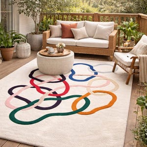 Può includere: Uno spazio abitativo all'aperto con un tappeto color crema con disegni a cerchio astratti e colorati. Il tappeto è posizionato su un ponte di legno, con mobili in vimini e piante in vaso. Un divano, una sedia e un tavolino completano la scena.