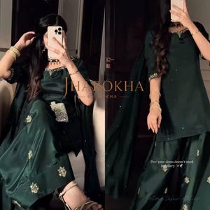 Puede incluir: Conjunto de satén verde esmeralda que incluye una blusa, pantalones anchos y una bufanda a juego. El atuendo está adornado con bordados y adornos dorados. El texto "Jharokha by Kha" y "Fatima tahir atelier" es visible.