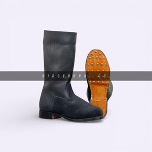 Puede incluir: Botas de cuero negro con empeine de ante y caña alta. Se ve la suela, con tachuelas metálicas y base de cuero marrón. Las botas se muestran sobre un fondo blanco.