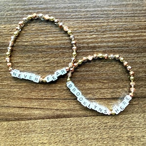 May include: Two beaded bracelets with gold and clear beads. One bracelet spells "NEVER FEAR" in white letter beads. The other bracelet spells "BROOKLYNS HERE" in white letter beads. The bracelets are on a wooden surface.
