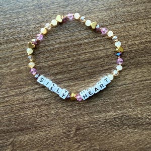 Könnte beinhalten: Ein Perlenarmband mit den Worten "SILLY HEART" aus weißen Buchstabenperlen. Das Armband hat goldene herzförmige Perlen, klare und lila Perlen sowie goldfarbene Abstandshalter. Das Armband liegt auf einer Holzoberfläche.
