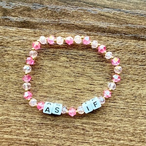 May include: A pink and clear beaded bracelet with the letters "ASIF" spelled out in white letter beads. The bracelet is made of small, faceted beads in shades of pink and clear, strung together on a flexible cord.