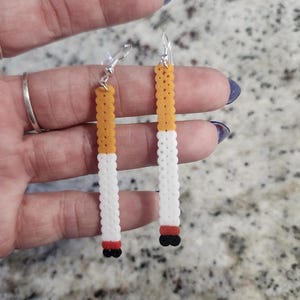 Op de afbeelding: Een paar novelty oorbellen ontworpen om op sigaretten te lijken. Elke oorbel heeft een witte, cilindrische behuizing met een geel-oranje filter en een rode en zwarte punt. De oorbellen hangen aan zilverkleurige oorhaakjes.