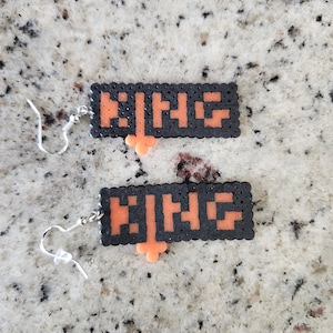 May include: A pair of black and orange pixelated earrings. Each earring features the word "KING" in orange pixelated letters on a black background. The earrings have silver-colored fish hook ear wires.