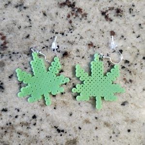 Op de afbeelding: Een paar lichtgroene, bladvormige oorbellen. Elke oorbel is gemaakt van kleine, ronde kralen en heeft een zilverkleurige haak. De oorbellen zijn ontworpen om op een cannabisblad te lijken.