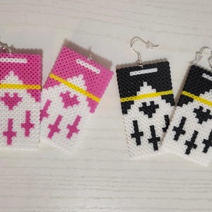 Op de afbeelding: Twee paar handgemaakte oorbellen. Eén paar is roze, wit en geel, de andere is zwart, wit en geel. De oorbellen zijn rechthoekig met een geometrisch ontwerp, gemaakt van kleine, gesmolten kralen.