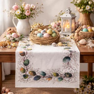 Puede incluir: Una mesa de Pascua está decorada con un camino de mesa blanco con un diseño botánico y huevos coloridos. Un nido de huevos se encuentra en el centro, rodeado de flores, una linterna y un conejito decorativo. La escena es brillante y festiva.