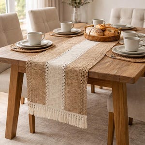 Op de afbeelding: Een eettafel gedekt voor een maaltijd, met een geweven tafelloper van jute met een centrale witte streep en franjes. De tafel is gedekt met borden, kopjes en placemats in neutrale tinten. Een mand met brood en broodjes staat in het midden.