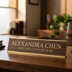 Könnte beinhalten: Ein Namensschild aus Holz mit goldfarbener Aufschrift „ALEXANDRA CHEN, CHIEF EXECUTIVE OFFICER“. Ein schwarzer Stift und ein geschlossenes Notizbuch liegen auf dem Schreibtisch. Eine Tasse Kaffee steht daneben.