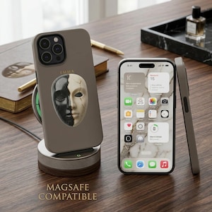 Peut inclure: Coque de téléphone taupe avec un motif de masque noir et blanc, présentée sur un chargeur sans fil. Un téléphone avec un écran à motif marbré est à côté. Le nom "EMMA" est imprimé sur la coque.