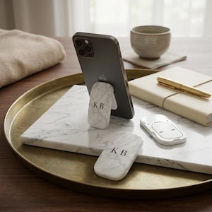 May include: A smartphone with a marble-patterned phone grip, displaying the initials "KB". The phone rests on a white marble surface, alongside a second grip and a white phone stand. A gold tray and notebook are also visible.