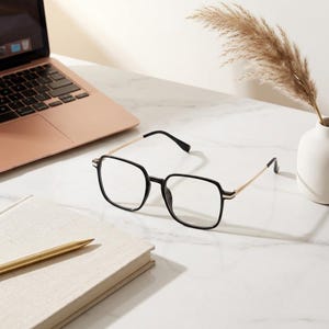 Può includere: Occhiali con montatura quadrata nera e aste sottili in metallo dorato. Gli occhiali presentano lenti trasparenti e sono posizionati su una superficie vicino a un portatile, un taccuino con una penna dorata e un vaso con erba della pampa.