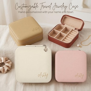 Op de afbeelding: Vier reissieradenkoffers in beige, crème en roze, waarvan één open om compartimenten te onthullen. Elke koffer is gepersonaliseerd met de naam "Ashley" in gouden script. De tekst "Customizable Travel Jewelry Case" staat bovenaan.