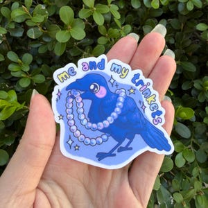May include: A sticker featuring a blue cartoon crow holding a pearl necklace. The text "me and my trinkets" is written around the crow. The sticker has a light blue background with star and sparkle accents.