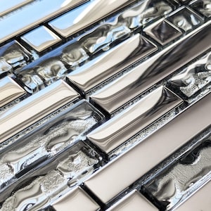 May include: Close-up of a mosaic tile surface. The tiles are a mix of rectangular and square shapes, with a reflective, metallic finish in shades of silver and grey. Some tiles have a textured, rippled appearance.