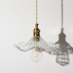 Peut inclure: Suspension luminaire en verre transparent avec un abat-jour en forme de fleur et un raccord en laiton. L'ampoule est visible à l'intérieur de l'abat-jour. Le cordon est torsadé beige.