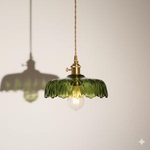 May include: A green glass pendant light with a scalloped shade and brass hardware. The light bulb is visible inside the shade. The cord is a twisted, light brown color. The light casts a shadow on the wall.