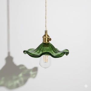 Peut inclure: Suspension en verre vert avec un abat-jour en forme de fleur froissée. Le luminaire a une douille de couleur laiton et une ampoule Edison visible. Le cordon est de couleur crème. La lumière est suspendue sur un fond blanc.