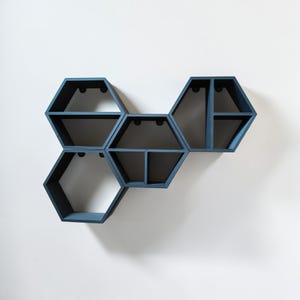 Könnte beinhalten: Ein Set aus fünf blauen sechseckigen Regalen, die an einer weißen Wand befestigt sind. Die Regale, mit unterschiedlichen Innenfächern, sind aus Holz gefertigt und in einem matten Blau lackiert. Das geometrische Design schafft einen modernen Look.