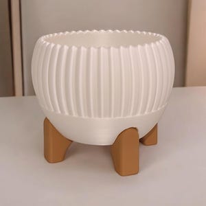 Könnte beinhalten: Ein weißer, gerippter Blumentopf mit gewelltem Rand, der auf drei braunen, abgewinkelten Beinen steht. Der kugelförmige Topf ist für Zimmerpflanzen konzipiert. Die neutrale Farbpalette eignet sich für verschiedene Einrichtungsstile.
