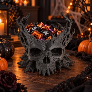 Könnte beinhalten: Eine dunkelgraue, texturierte Schale in Totenkopfform mit gezackten, baumartigen Aststrukturen, die nach oben ragen. Die Schale ist mit verschiedenen kleinen, einzeln verpackten Bonbons gefüllt, die mit orangefarbenen Kürbis- und weißen Geistermotiven bedruckt sind.