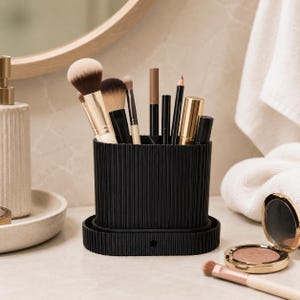 Könnte beinhalten: Ein schwarzer, geriffelter Make-up-Pinselhalter, gefüllt mit verschiedenen Pinseln, Stiften und Tuben. Der Halter steht auf einem passenden Sockel, neben einem Kompaktpuder und einem Make-up-Pinsel. Der Hintergrund umfasst einen Spiegel und ein weißes Handtuch.