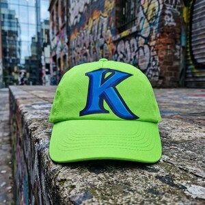 May include: A bright green baseball cap with a large blue letter 'K' outlined in white. The cap is made of a textured material and has a curved peak. The background shows a brick wall covered in colourful graffiti.