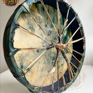 Puede incluir: Un tambor redondo hecho a mano con un parche de piel natural y un marco verde azulado oscuro. El parche del tambor está estirado sobre un marco de madera y asegurado con cordones de cuero. El logotipo "ZAMJ Crafts Drums & Musical Instruments" es visible.