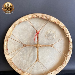 May include: A round, handcrafted drum with a natural hide surface and a wooden frame. The drum features a central wooden cross design with attached strings and a red ribbon. The drum's frame is tan, and the hide is a light beige color.