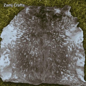 May include: A cowhide rug featuring a mottled pattern of dark brown and white fur. The hide has an irregular, organic shape with a dense, textured coat. The text Zamj Crafts is printed in the upper left corner of the image.