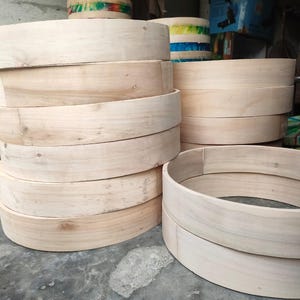 Puede incluir: Una pila de aros de madera de color claro sin terminar con un diseño circular liso. Estas bandas circulares están hechas de madera natural, mostrando patrones de grano visibles y un tono beige claro, dispuestas en varias pilas verticales.