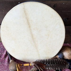 May include: A circular frame drum with a light beige, textured skin head featuring a subtle diagonal line across the surface. The drum is accompanied by a wooden mallet with a wrapped head and a decorative feather accent with brown and black stripes.