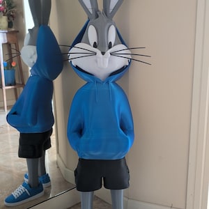 Puede incluir: Una gran estatua gris de Bugs Bunny con una sudadera con capucha azul, pantalones cortos negros y zapatillas azules con cordones blancos. La estatua tiene largas orejas grises y bigotes negros. La figura está frente a un espejo.