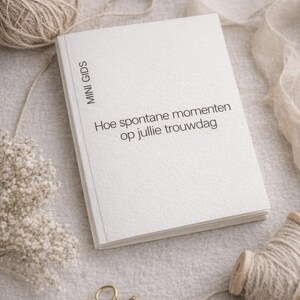 Puede incluir: Un libro blanco con el texto "Hoe spontane momenten op jullie trouwdag" y "MINI GIDS" impreso en la portada. El libro está sobre una superficie de color neutro con una bobina de hilo, un clip dorado y flores decorativas.